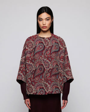 A woman stands against a plain background wearing the MIRTO 1956 Burgundy Paisley Jacquard Jacket—an oversized, cotton-blend piece in rich red and purple tones from MIRTO 1956—styled over a long burgundy dress and black shoes. The relaxed fit of the jacket complements her straight dark hair and neutral expression.