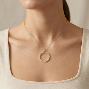 A woman wearing a cream square-neck top and the ELLA STEIN - YOU ARE MY EVERYTHING PENDANT NECKLACE by ELLA STEIN, featuring a large, circular, diamond-studded pendant. The natural diamond necklace adds an elegant touch to her look, complemented by her neatly tied-back hair and the neutral background.