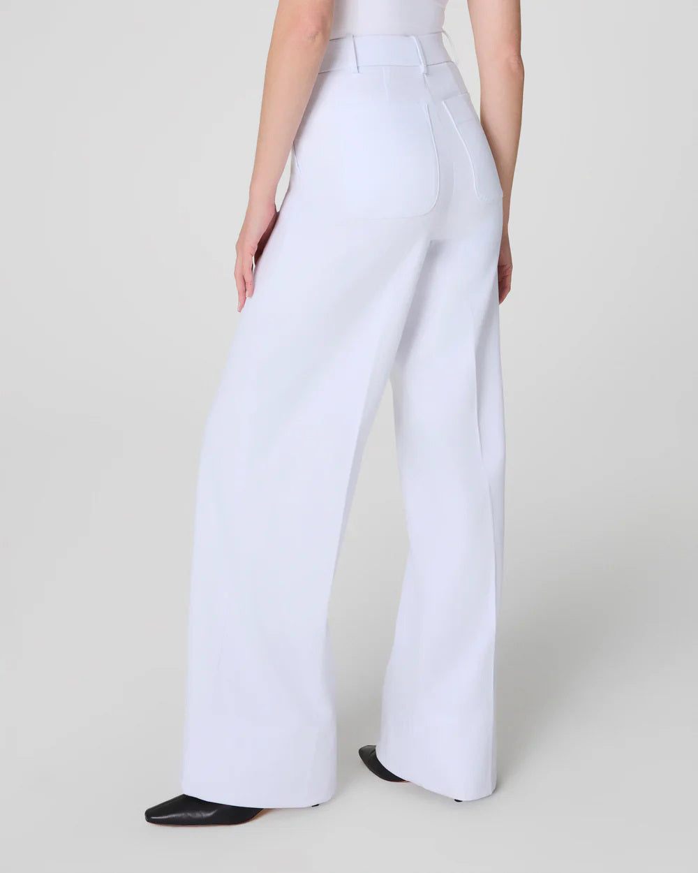 A woman with curly hair stands against a stucco wall, embodying minimalistic elegance in SPANX Stretch Twill Wide Leg Pant and a white sleeveless top. She poses with a black handbag and hand in her pocket, showcasing chic style and 360-degree smoothing comfort.