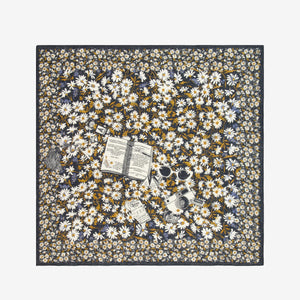 A woman with short hair, in a black sleeveless top and blue pants, stands against a white background. She wears the INOUI EDITIONS FAUST SCARF IN BLACK from INOUI, featuring yellow and white flowers, wrapped around her neck.