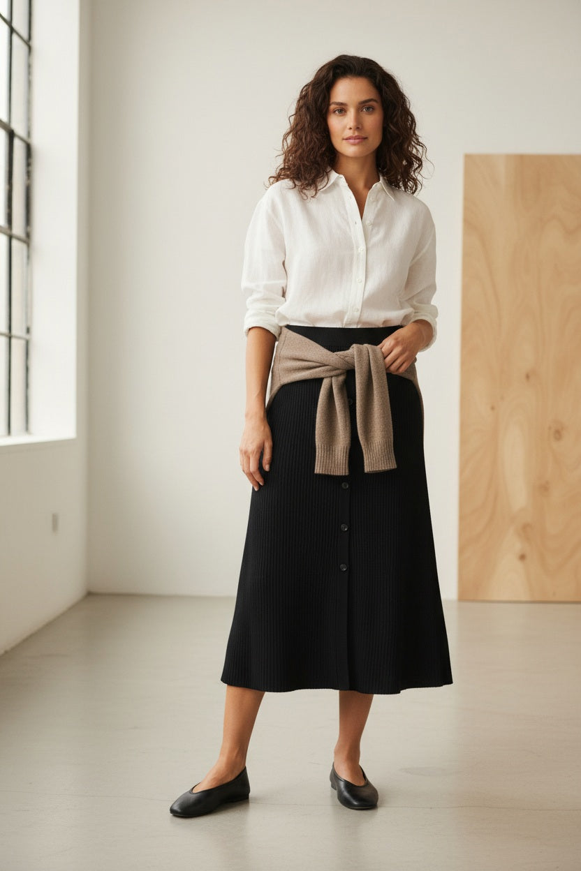 A woman with wavy brown hair stands in a sunlit room, smiling as sunlight streams through large windows. She wears a white button-up shirt paired with the HARTFORD - MELKA KNIT SKIRT IN BLACK by HARTFORD, black flats, and a gold bracelet.