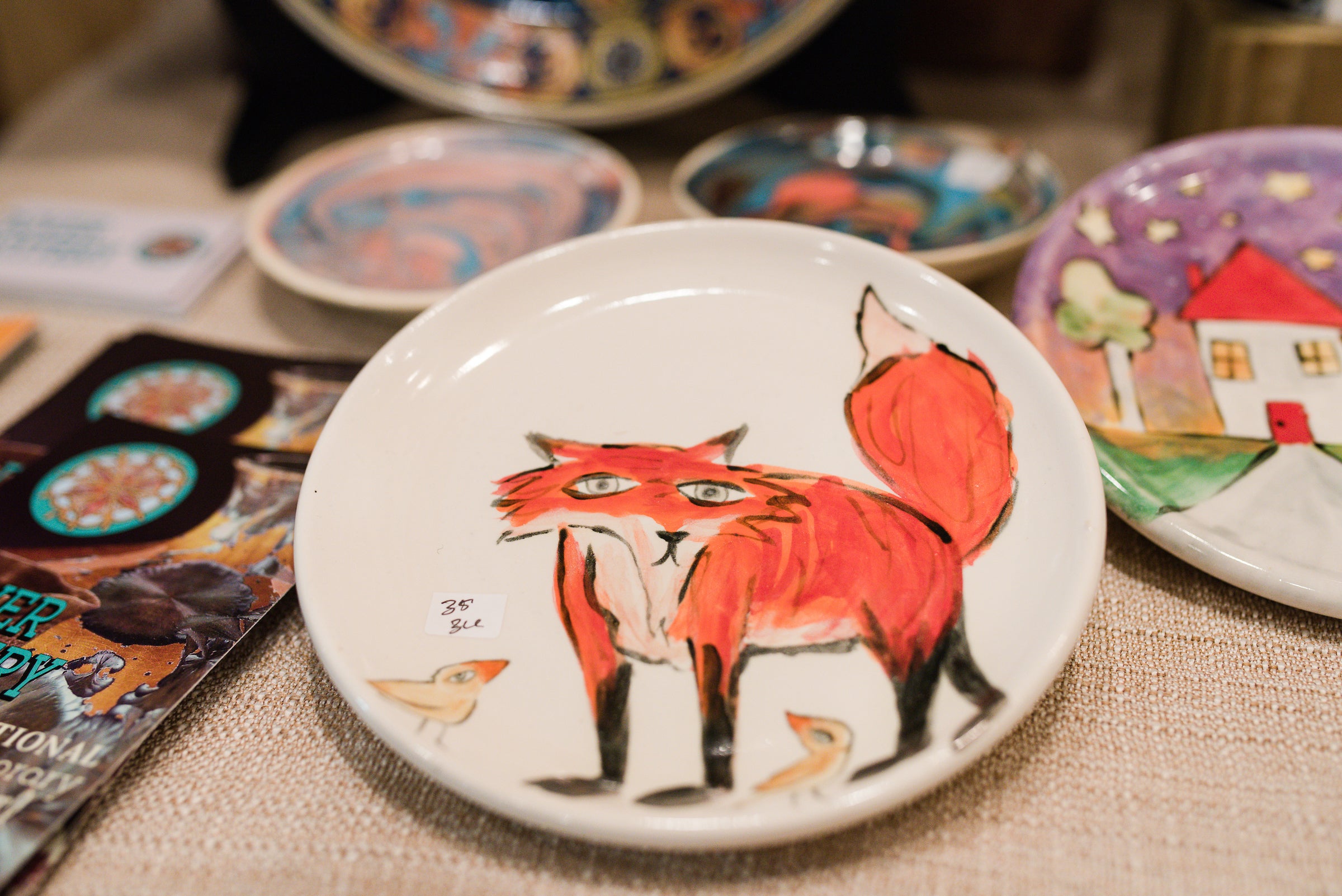 A ceramic plate featuring a hand-painted red fox with two small birds. The plate is displayed on a table among other colorful ceramic dishes with various designs. A price tag is visible on the plate.