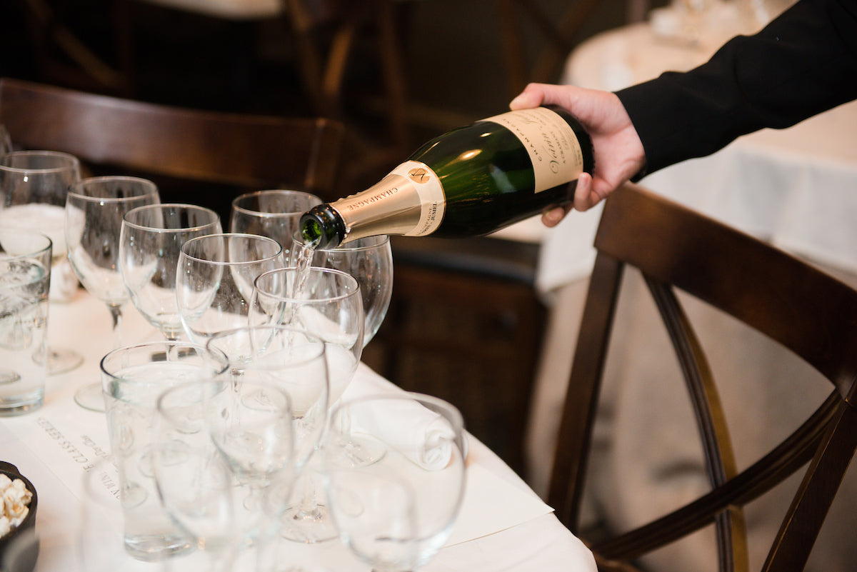 A person pours champagne from a bottle into several empty wine glasses arranged on a white tablecloth at a dining table set for an event.