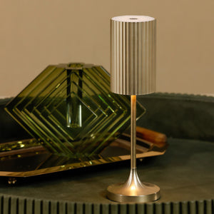 The ZODAX REED RECHARGEABLE LAMP is a modern, gold-colored table lamp with a cylindrical, vertically ribbed shade and a sleek, straight stem on a round base. It features adjustable light intensity and is shown against a white background.