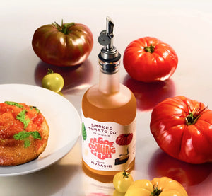 A glass bottle labeled “ALGAE COOKING CLUB - MUSASHI’S SMOKED TOMATO OIL” featuring illustrations of tomato and basil, with a wooden cap, contains amber-colored oil. Produced by ALGAE COOKING CLUB, this oil is ideal for adding a rich, savory grilled tomato flavor to your dishes.