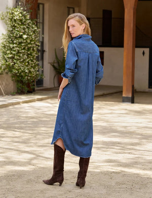 A woman stands outdoors in the FRANK & EILEEN Rory Maxi Shirtdress in Distressed Indigo Washed Denim, its long button-up silhouette and rolled sleeves crafted from luxurious indigo fabric. She pairs the effortlessly chic dress with brown knee-high boots, set against a backdrop of lush greenery and a building.