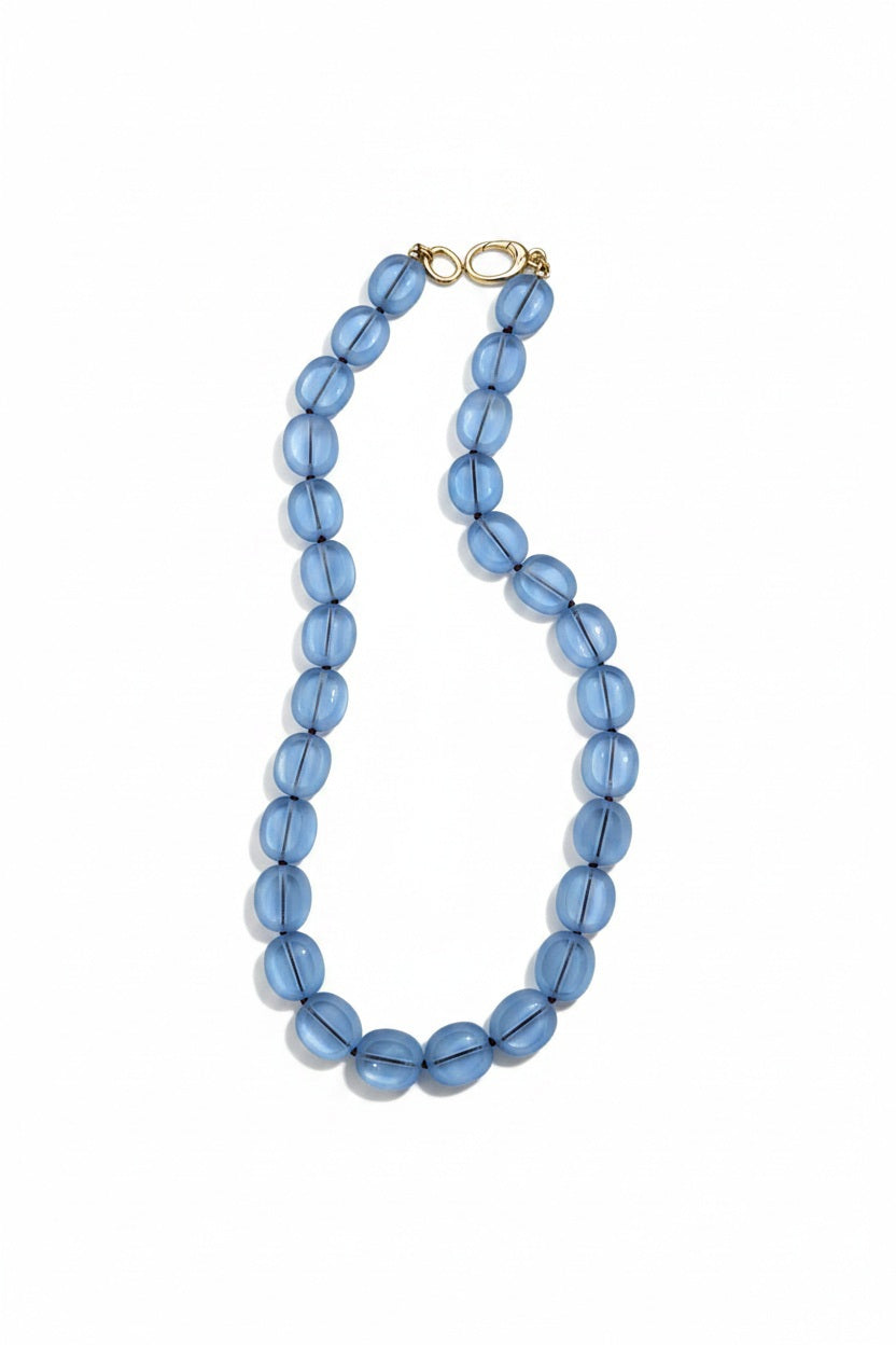The SANDRINE FRANCE STUDIO - STEFANIE NECKLACE IN AQUA BURGUNDY by SANDRINE FRANCE STUDIO is a handmade necklace showcasing large, translucent blue oval beads paired with a gold plated clip, elegantly displayed on a white background.