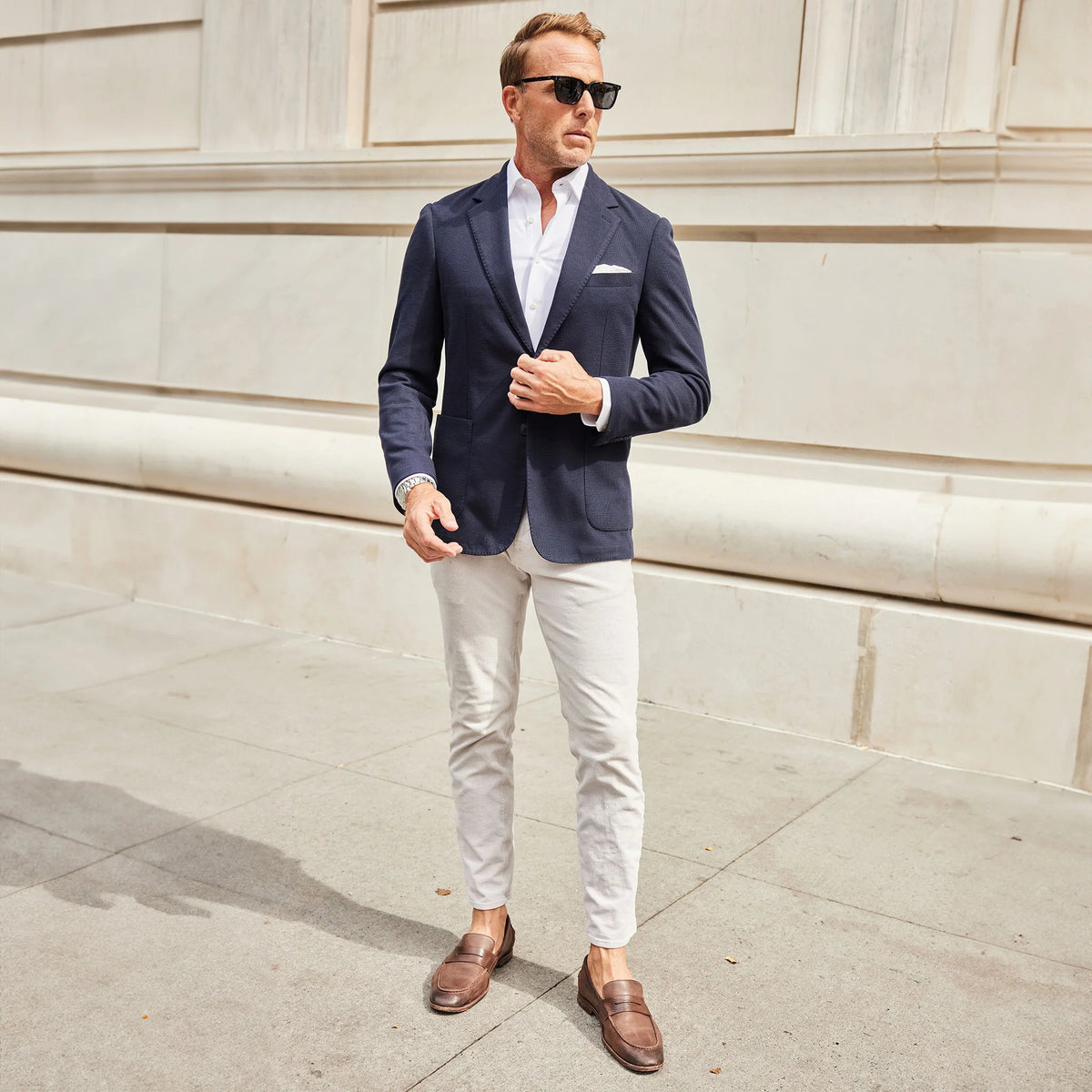 A man in the COLLARS AND CO Maverick Knit Unstructured Blazer in navy, paired with a light blue shirt and sunglasses, stands on a busy city street with cars, bikes, and buildings behind him.