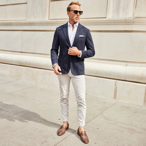 A man in the COLLARS AND CO Maverick Knit Unstructured Blazer in navy, paired with a light blue shirt and sunglasses, stands on a busy city street with cars, bikes, and buildings behind him.