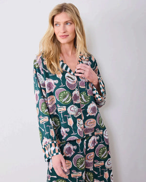 A woman stands barefoot on a wooden floor against a white wall, wearing the PRINTFRESH - Champagne & Caviar Eco Satin Soho Set in Peacock Feather by PRINTFRESH, a matching dark loungewear set adorned with colorful floral and fruit motifs and finished with checkered cuffs.