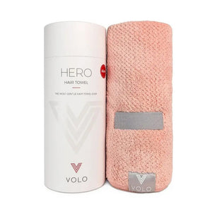 A person with short dark hair, wearing a white shirt, wraps their hair in the VOLO BEAUTY - HERO HAIR TOWEL LUXE and adjusts it with both hands in a bright, airy space filled with soft natural light.