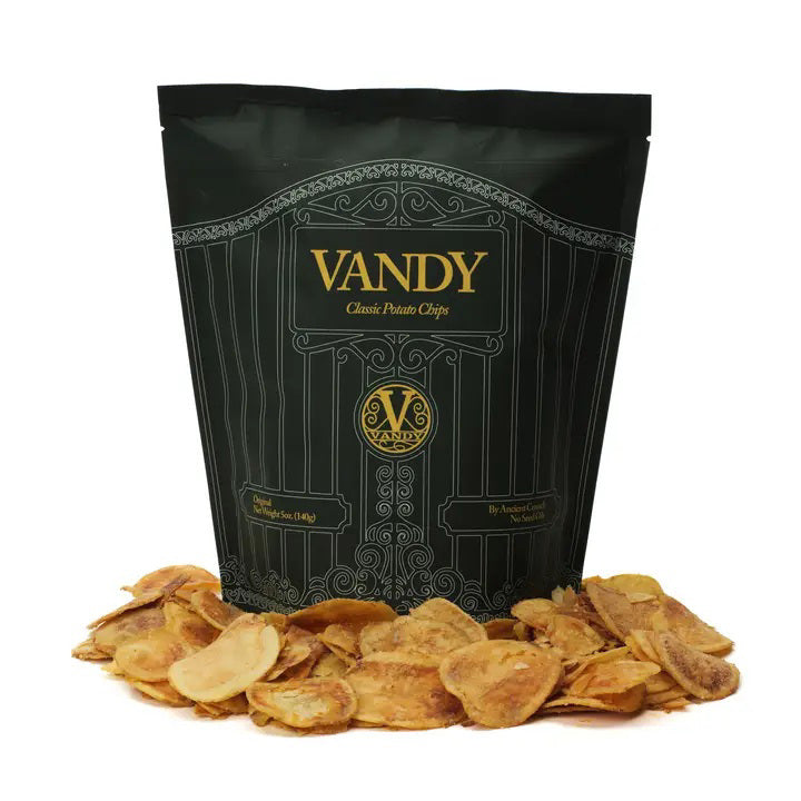 A black bag labeled &quot;ANCIENT CRUNCH - VANDY TRADITIONAL POTATO CHIPS&quot; stands upright with a stylish design, surrounded by a scattered pile of golden-brown potato chips in front of it.