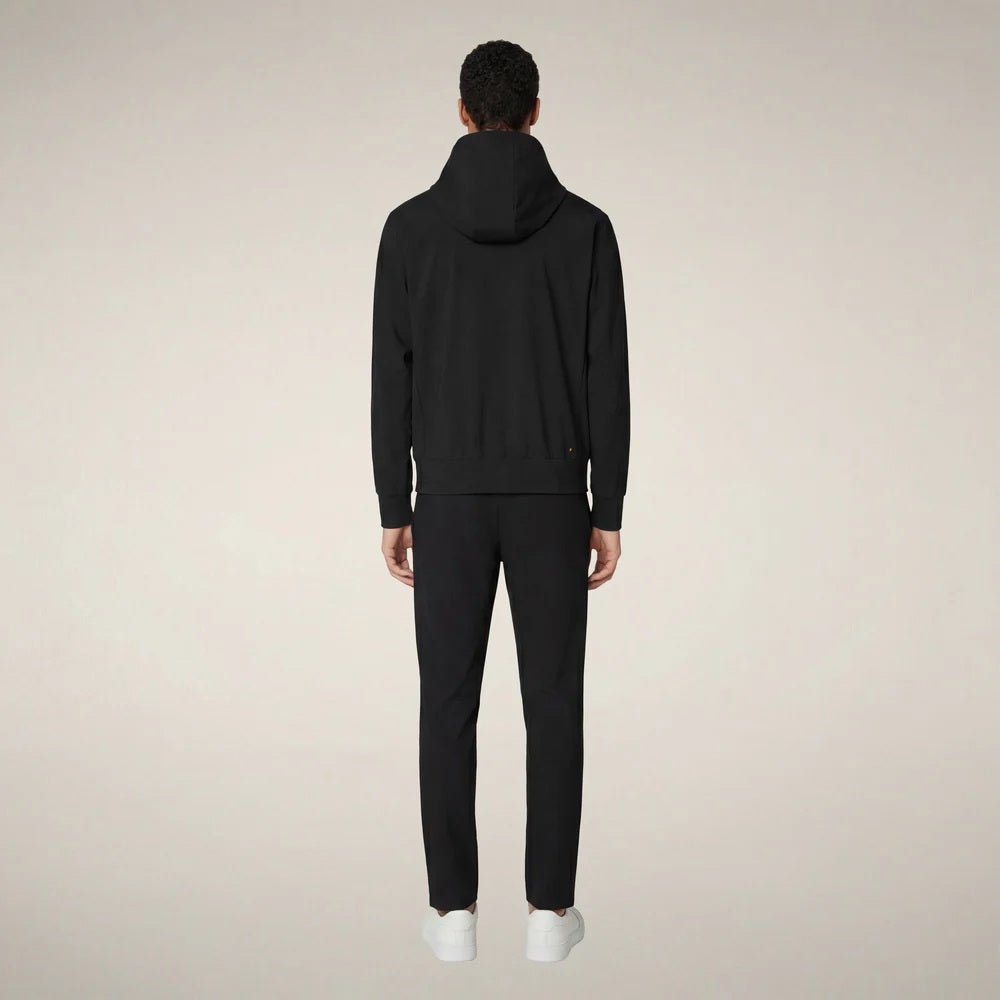 A man faces forward in the SAVE THE DUCK - LUIZ Hooded Jacket in Blue Black from SAVE THE DUCK, paired with black pants and white sneakers, set against a plain beige background.