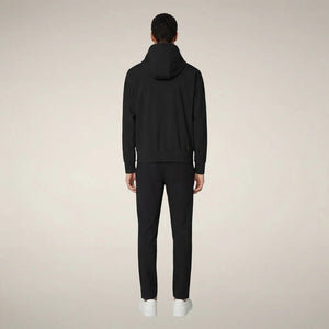 A man faces forward in the SAVE THE DUCK - LUIZ Hooded Jacket in Blue Black from SAVE THE DUCK, paired with black pants and white sneakers, set against a plain beige background.