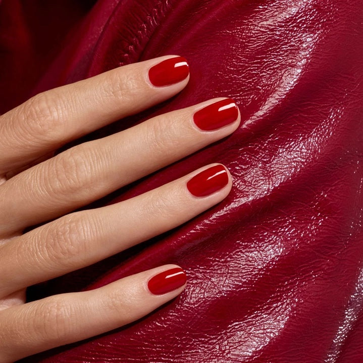 A hand with neatly manicured nails painted in MANUCURIST - GREEN™ NAIL POLISH IN RED VELVET by MANUCURIST INC rests on a textured red leather surface, creating a coordinated and stylish look.