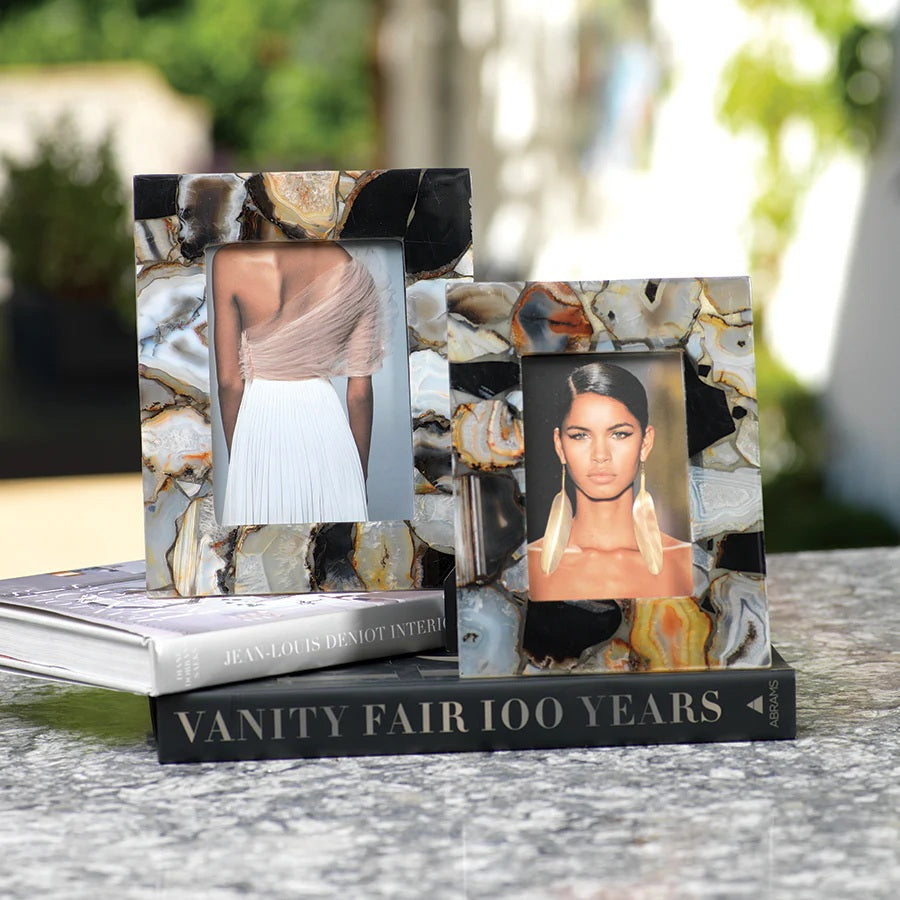 Two decorative photo frames, including the ZODAX BAHIA AGATE FRAME - 4X6, sit on stacked books outdoors; one frame displays a woman’s back in a light top, while the other shows a woman with earrings facing forward. The top book is "JEAN-LOUIS DENIOT INTERIORS.