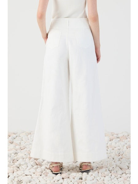 A person stands on white pebbles, shown from the back, wearing ELEH’s WIDE LEG COTTON TROUSER paired with a white sleeveless top and sandals—an effortless take on modern minimalism.