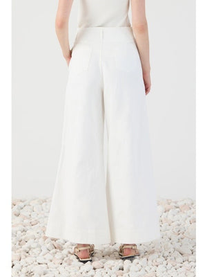 A person stands on white pebbles, shown from the back, wearing ELEH’s WIDE LEG COTTON TROUSER paired with a white sleeveless top and sandals—an effortless take on modern minimalism.