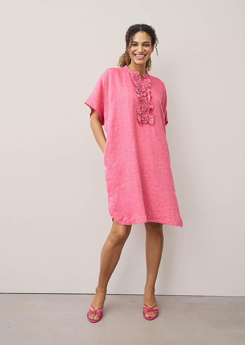 A woman models the 0039 ITALY DAISY LINEN DRESS, featuring ruffled detailing along the button placket. Only her lower face and upper torso are visible as she stands against a light background, showcasing modern femininity.