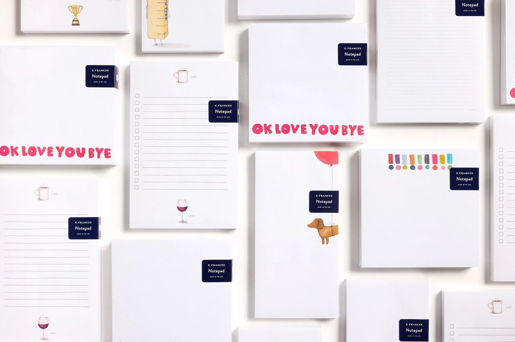 The E. FRANCES - NOTEPAD HOTDOG HIGH by E. FRANCES PAPER features a brown dachshund tied to a red heart balloon on a white background—ideal for pet lovers&#39; stationery and proudly made in the USA.