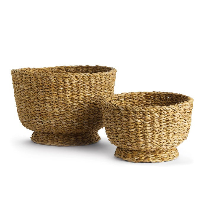 The NAPA HOME AND GARDEN Seagrass Footed Bowls feature two round, natural light brown woven bowls, each with a wide opening and flared base—ideal for natural home decor. The larger bowl sits behind and left of the smaller one.
