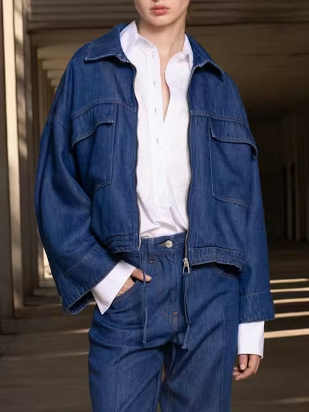 A person in a loose-fitting NUDE - DENIM JACKET by NUDE and matching jeans is seen from behind, standing in a shaded outdoor area.