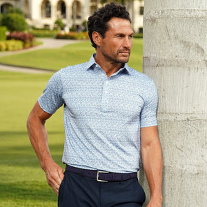 A man in the COLLARS AND CO Featherweight Flex Polo in Coastal Blue Floral with a dress collar and dark pants stands outdoors by a large pillar, with green grass, gardens, and a building in the background.