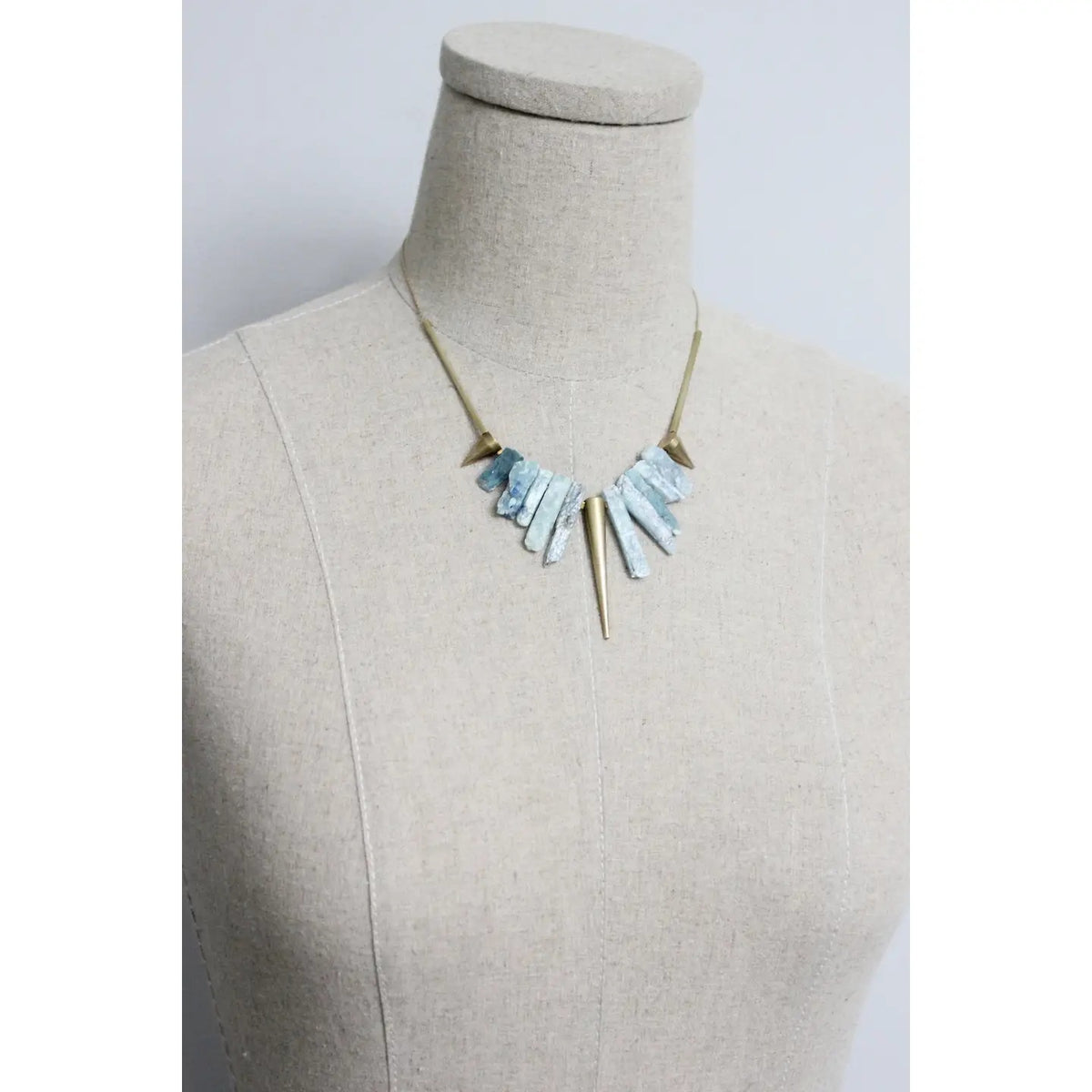 The DAVID AUBREY - KYANITE AND BRASS GEOMETRIC NECKLACE by DAVID AUBREY features a gold tone chain adorned with blue kyanite beads and geometric brass accents arranged symmetrically, creating a striking necklace effect against a gray background.
