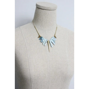 The DAVID AUBREY - KYANITE AND BRASS GEOMETRIC NECKLACE by DAVID AUBREY features a gold tone chain adorned with blue kyanite beads and geometric brass accents arranged symmetrically, creating a striking necklace effect against a gray background.