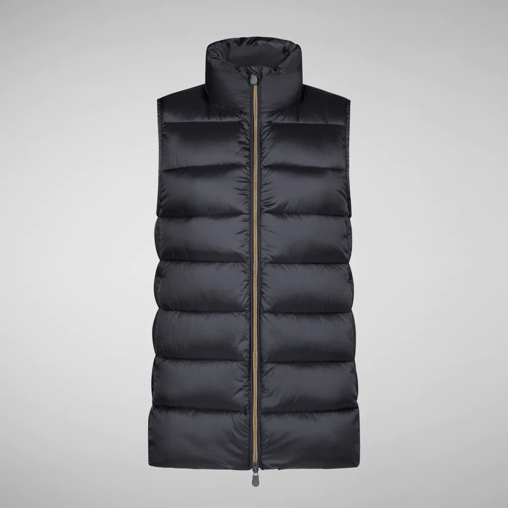 A person wearing a black cap, sunglasses, the SAVE THE DUCK - CORAL PUFFER VEST with animal-free down from SAVE THE DUCK over a black long-sleeve top, beige wide-legged pants, and black ankle boots stands against a plain background. The outfit appears to be styled for colder weather.