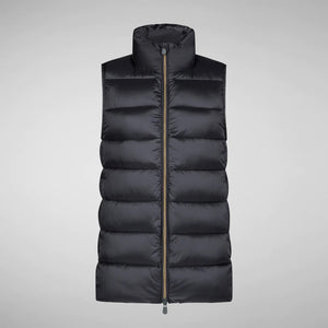 A person wearing a black cap, sunglasses, the SAVE THE DUCK - CORAL PUFFER VEST with animal-free down from SAVE THE DUCK over a black long-sleeve top, beige wide-legged pants, and black ankle boots stands against a plain background. The outfit appears to be styled for colder weather.