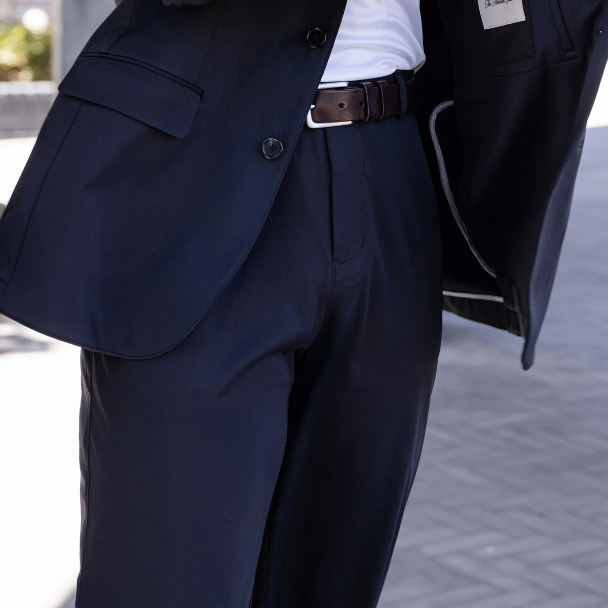 A man in a dark suit and COLLARS AND CO - MAVERICK PERFORMANCE PANTS IN BLACK, wearing a white shirt and sunglasses, leans casually against a gold wall with warmly lit columns in the background, holding a phone.