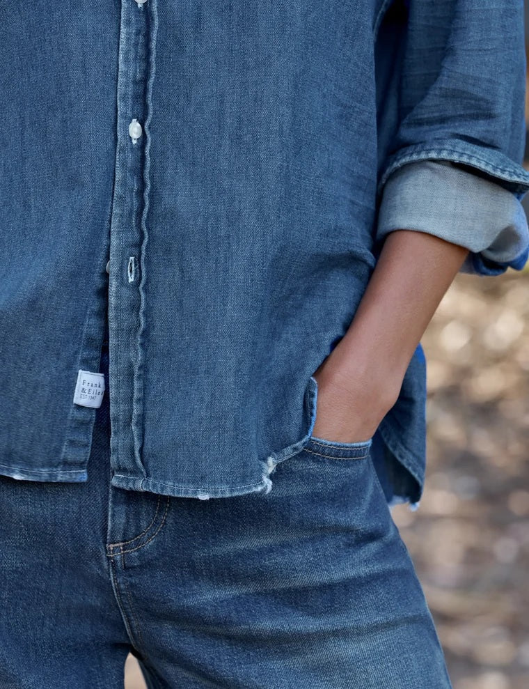 A person with long brown hair stands outdoors, facing away, next to a black wooden fence with trees and grass in the background, wearing the FRANK & EILEEN EILEEN Relaxed Button Up Shirt in Famous Denim Distressed Indigo Wash.