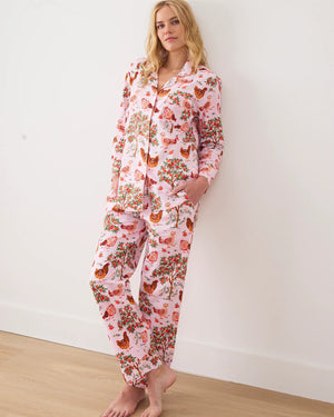 A person stands barefoot against a white wall in PRINTFRESH's pink organic cotton HEN HOUSE PAJAMA SET, featuring hand screen-printed roosters and floral patterns. The pajamas have long sleeves and pants, with one hand in a pocket and their head slightly tilted.