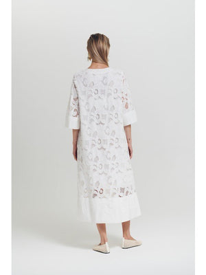 A woman wears the MILVA MI - EMBROIDERED COTTON DRESS IN WHITE by MILVA, styled with bold black and white earrings and black sunglasses. Her straight blonde hair and hand on her hip complete a chic summer resort look.
