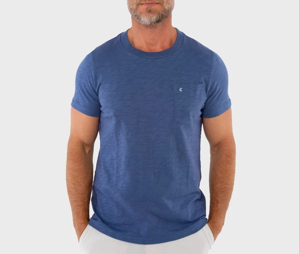 A man wearing the CRIQUET Slub Cotton Short Sleeve Pocket T-Shirt in Vintage Indigo stands against a textured white wall, paired with light-colored pants and one hand in his pocket. His face is partially out of frame.