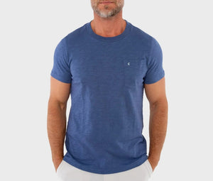 A man wearing the CRIQUET Slub Cotton Short Sleeve Pocket T-Shirt in Vintage Indigo stands against a textured white wall, paired with light-colored pants and one hand in his pocket. His face is partially out of frame.