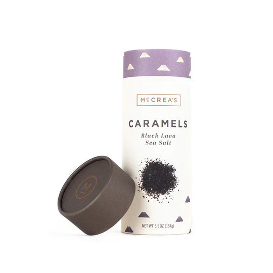 A cylindrical container of McCREA'S CANDIES Black Lava Sea Salt Caramels stands upright with its lid placed beside it. The container is adorned with a purple band featuring small triangular shapes, and the weight is listed as 5.5 oz (154 g).