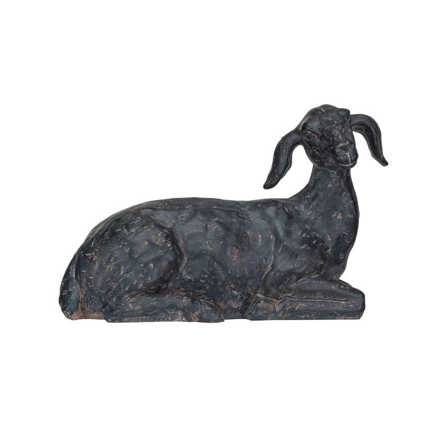 Two CREATIVE COOP RESIN GOAT WITH CAST IRON FINISH figurines are placed on a light-colored surface. One goat is sitting on a stack of books, while the other stands beside it. Small scattered grains or pebbles adorn the base of the standing goat. The background includes a plain, light wall, adding to this charming accent decor.