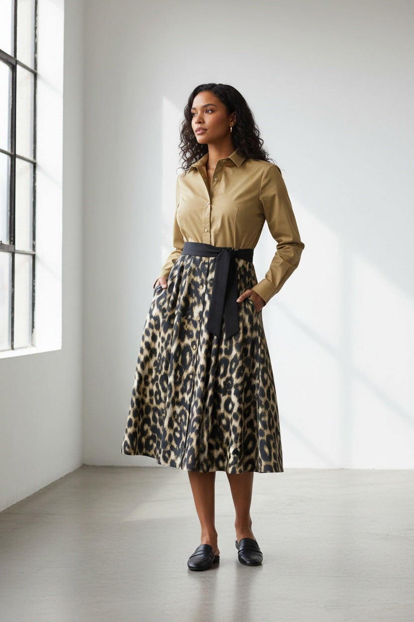 A woman stands facing forward wearing a tan long-sleeve button-up shirt tucked into a knee-length leopard print skirt with a black belt, against a plain light gray background.