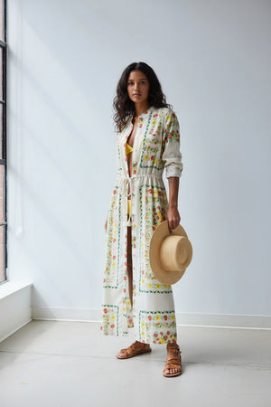 A person stands facing away in the HOPE FOR FLOWERS Band Collar Shirtdress in Custard Floral, paired with brown sandals and holding a straw hat, against a plain light gray background.