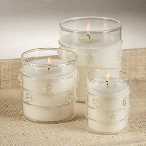 Two lit ZODAX Grand Casablanca Candle – Wild Tuberose, 6.5oz, in decorative glass holders rest on patterned boxes beside a frosted white pitcher with a green rim, all on a neutral surface, evoking the wild tuberose scent.