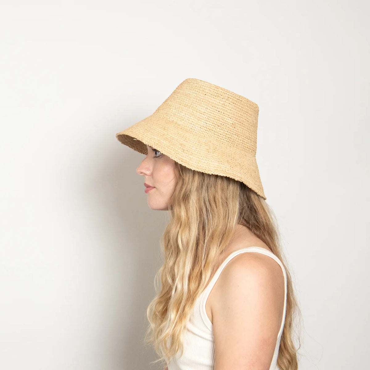 The WYETH HATS - TALI RAFIA BUCKET HAT IN NATURAL by WYETH is a wide-brimmed, natural straw UPF 50+ hat with a rounded crown and textured woven pattern, displayed on a plain white background.