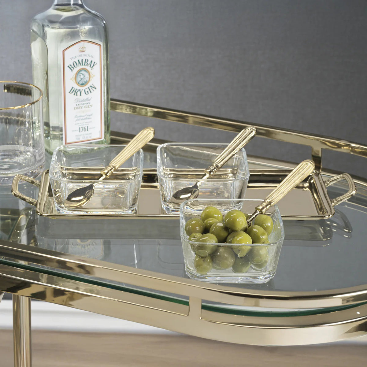 The ROUSSILLON CONDIMENT SET by ZODAX features three clear glass bowls with brass spoons, displayed on a rectangular gold metal tray with handles against a white background.
