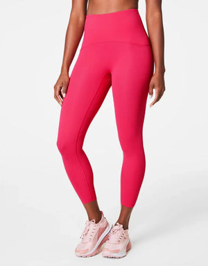 A person wears SPANX - BOOTY BOOST 7/8 LEGGINGS in high-waisted royal blue performance fabric with black athletic shoes, standing hand on hip against a plain white background. Only their lower body is visible; upper body and face are not shown.