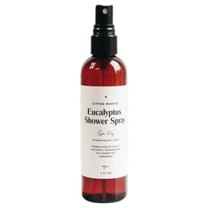 A hand holds a brown spray bottle labeled “SPA DAY EUCALYPTUS SHOWER SPRAY” by GIFTEN MARKET—an uplifting eucalyptus aromatherapy mist—set against a black showerhead and white marble shower wall, ideal for transforming your shower into a spa-like escape at home.