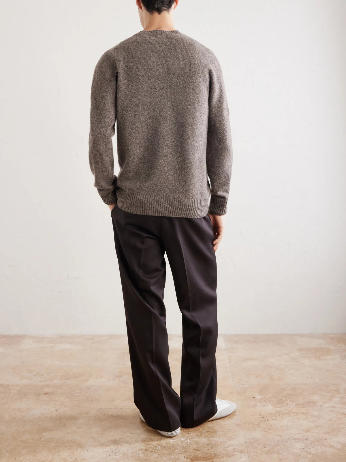 A person wearing the HARTFORD Seamless Wool Crew Pullover in Mushroom, an Italian-made sweater, paired with dark pants, is seated with one arm resting on their knee against a plain light background.