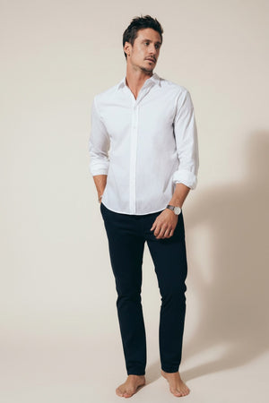 A man in the FRANK & EILEEN Finbar Men's Button Up Shirt in classic white poplin stands with rolled sleeves and hands in pockets, looking to the side. He has short dark hair, wears a watch, and poses against a light background.