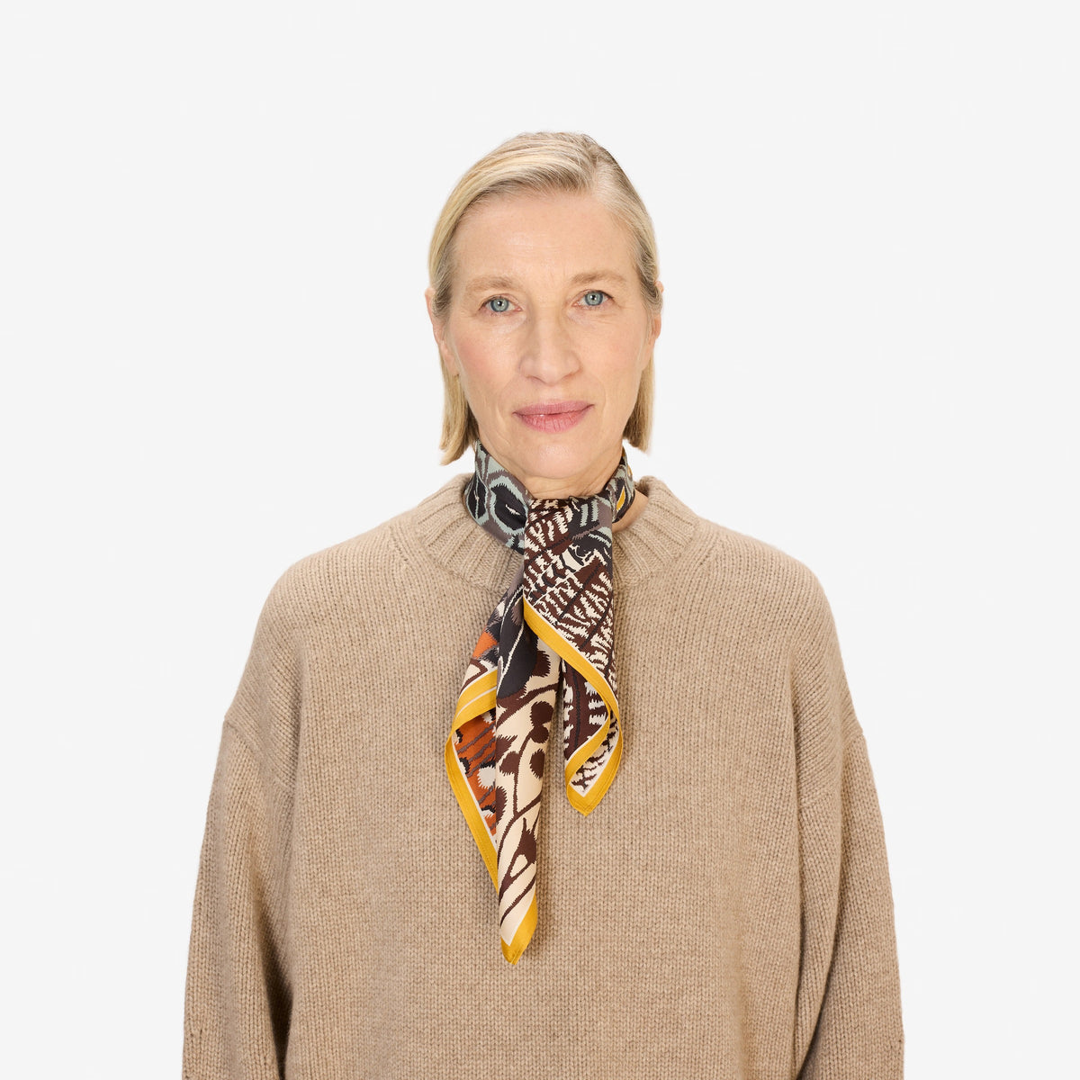 A close-up of a person wearing the INOUI EDITIONS - PAPILLON (BUTTERFLIES) SQUARE WOOL SCARF IN NATURAL by INOUI, draped around their neck over a black top. The scarf showcases abstract and geometric butterfly designs in earthy tones.
