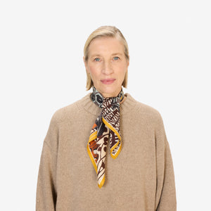A close-up of a person wearing the INOUI EDITIONS - PAPILLON (BUTTERFLIES) SQUARE WOOL SCARF IN NATURAL by INOUI, draped around their neck over a black top. The scarf showcases abstract and geometric butterfly designs in earthy tones.